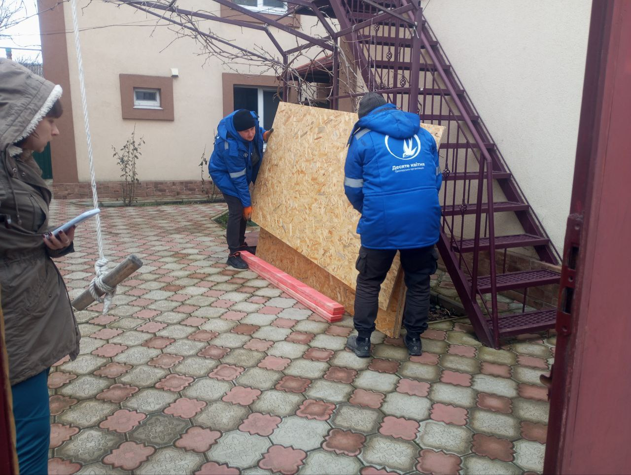 Двоє чоловіків у синій формі з логотипом благодійної організації «Десяте квітня» розвантажують листи OSB-плит на подвір'ї приватного будинку. Поруч стоїть жінка, яка робить запита у блокноті. На фоні видно металеві сходи на другий поверх будівлі.