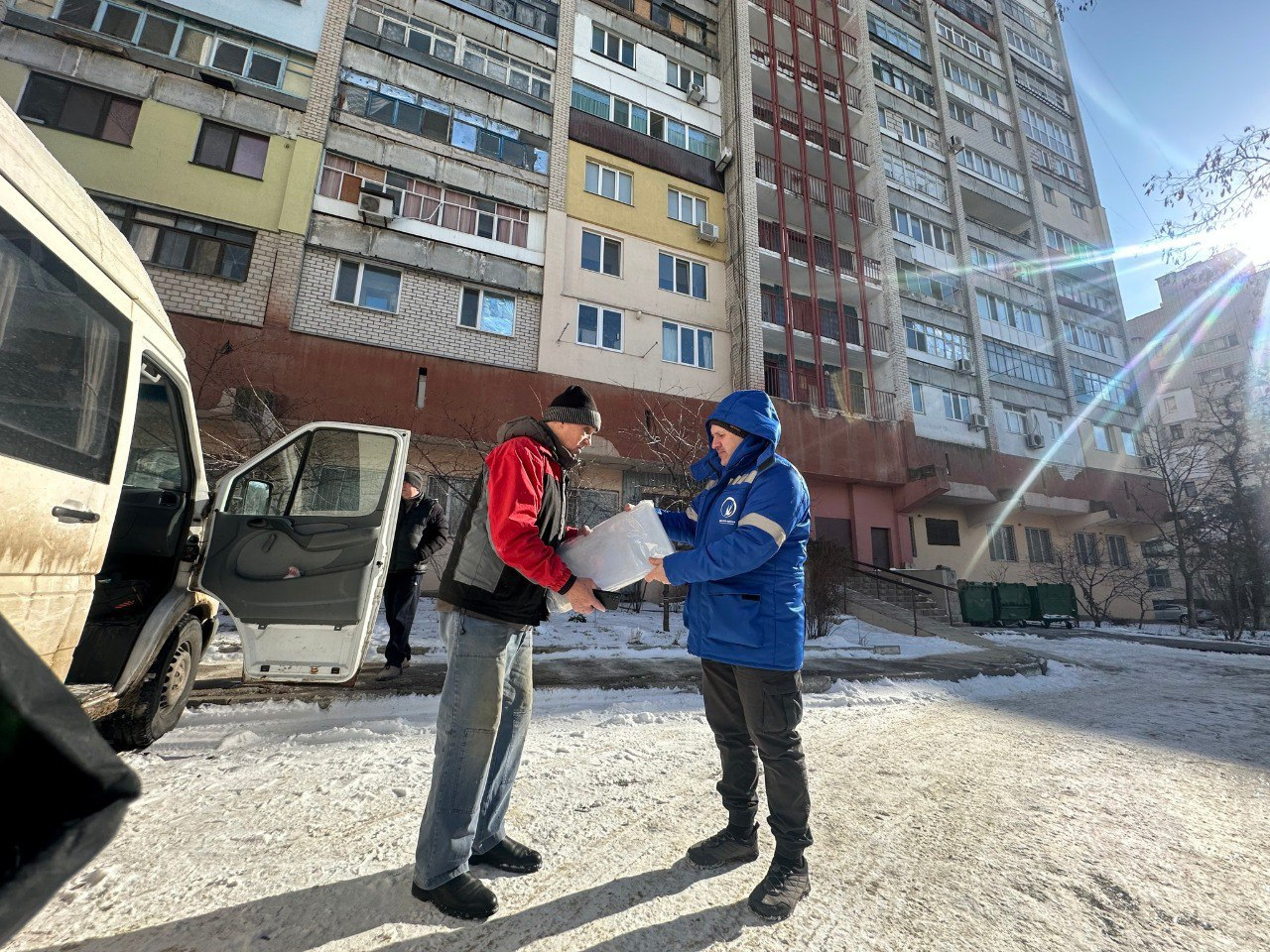 На засніженому подвір'ї біля мікроавтобуса волонтер у синій куртці передає чоловікові пакунок із гуманітарною допомогою. На задньому плані видно багатоповерховий житловий будинок у сонячний день.