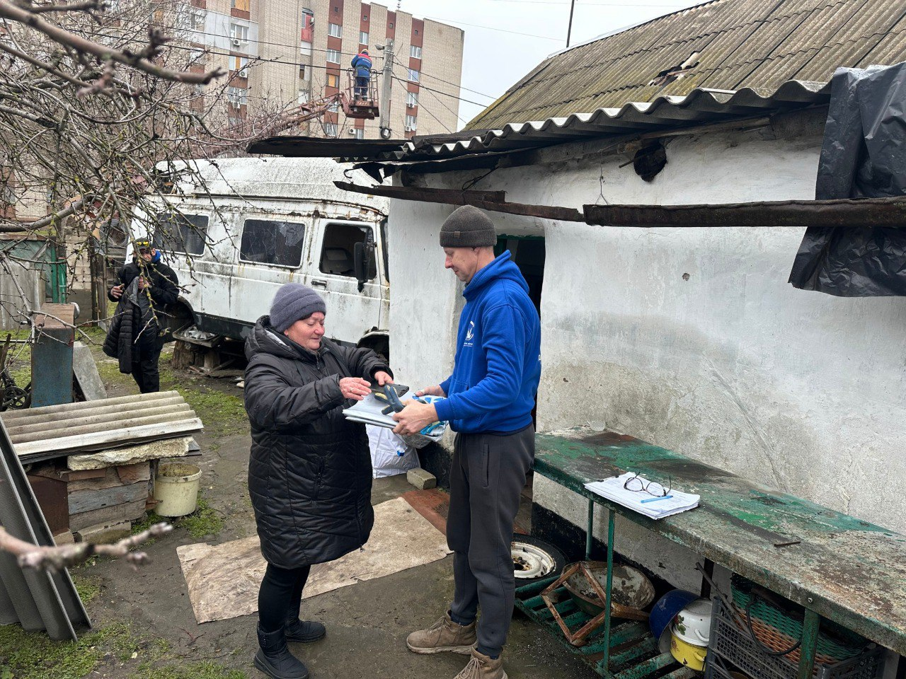 На подвір'ї біля невеликої білої будівлі чоловік у синій толстовці передає жінці пакунок із гуманітарною допомогою. На задньому плані видно старий білий фургон та багатоповерховий житловий будинок.