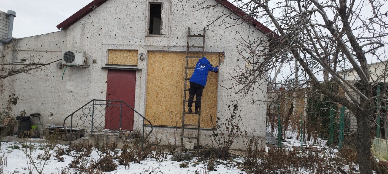 Людина в синій куртці стоїть на драбині та забиває вікно пошкодженого будинку OSB-плитою. Стіни будівлі посічені уламками, а земля навколо вкрита залишками снігу.