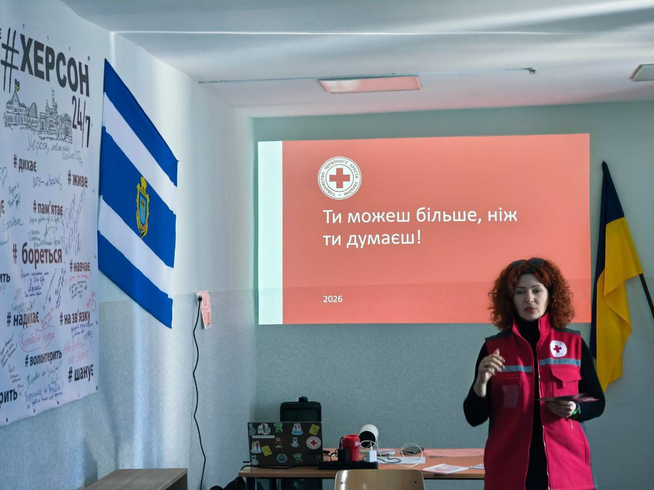 Представниця Червоного Хреста проводить презентацію в аудиторії. Позаду неї — екран із написом «Ти можеш більше, ніж ти думаєш!», державний прапор України та прапор Херсона.