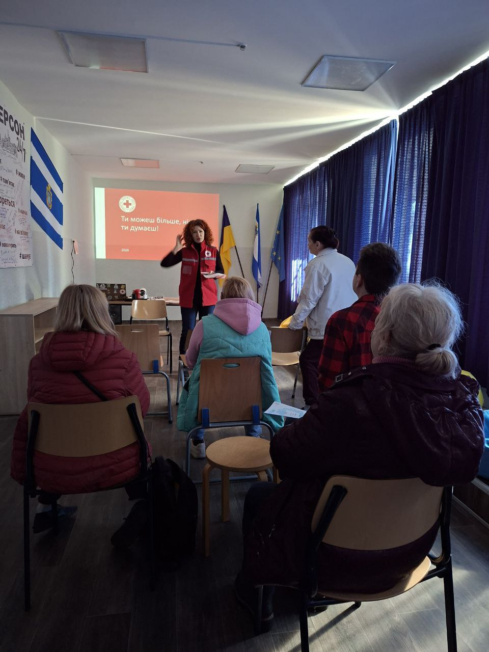 Жінка у жилеті Червоного Хреста проводить презентацію в аудиторії перед слухачами. На фоні — проєкція з написом «Ти можеш більше, ніж ти думаєш!» та прапори України і Херсона.