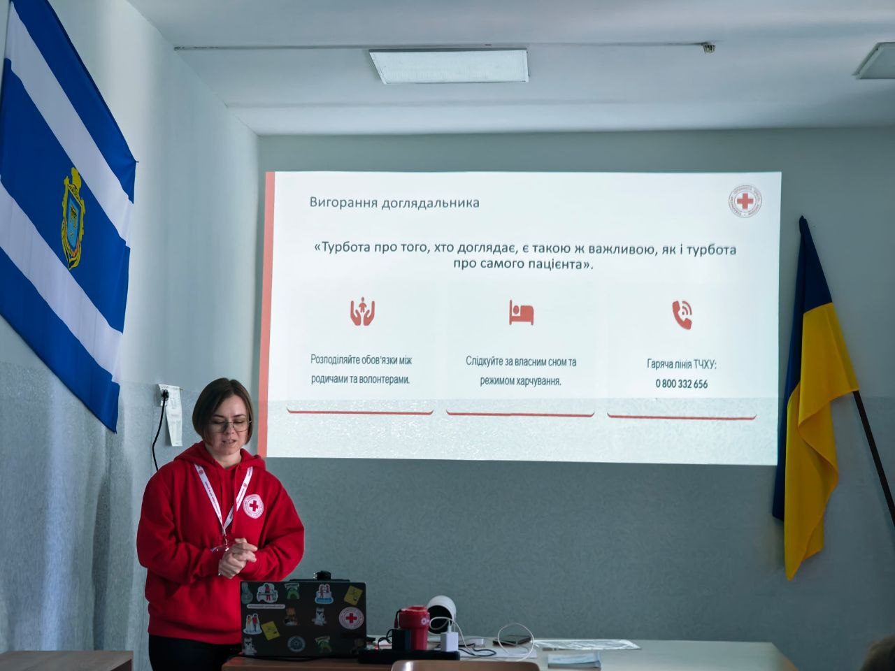 Жінка в червоному худі з емблемою Червоного Хреста проводить презентацію про вигорання доглядальників. Позаду неї — екран зі слайдом та прапори України і Херсона.