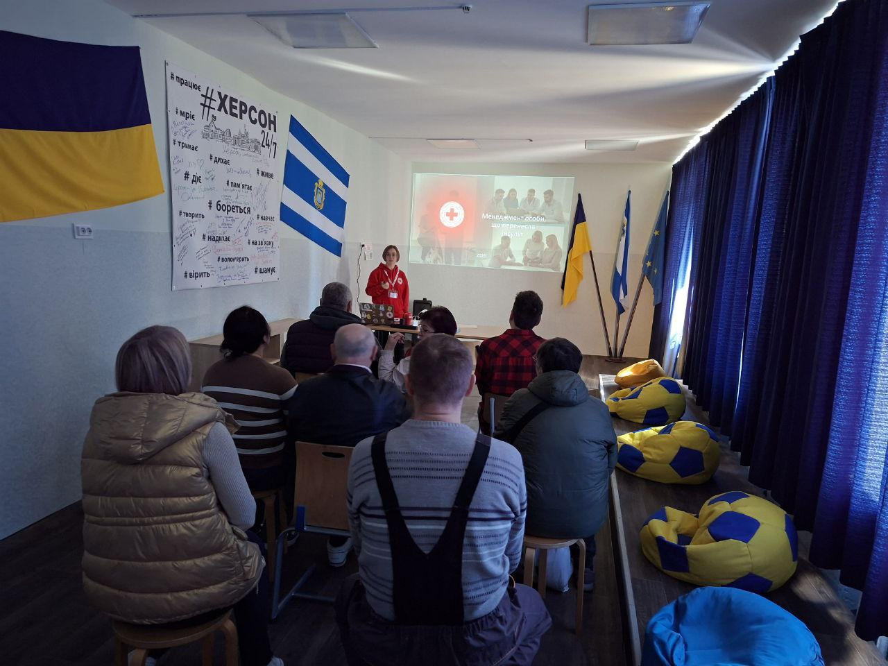 Спікерка в червоному худі з емблемою Червоного Хреста проводить презентацію в аудиторії перед групою людей. У приміщенні видно прапори України та Херсона, плакат із патріотичними написами та крісла-мішки.
