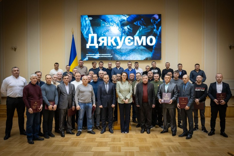 Групове фото чоловіків та жінок в офіційному залі на фоні екрана з написом «Дякуємо». Дехто тримає в руках нагороди. Позаду стоїть державний прапор України.