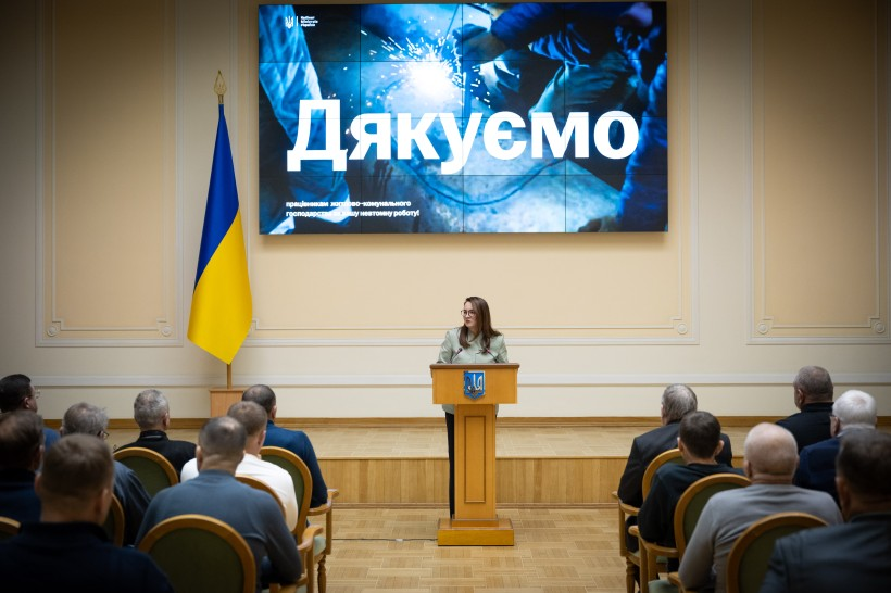 Жінка виступає за трибуною з гербом України в офіційній залі перед аудиторією. На великому екрані позаду неї напис «Дякуємо», а ліворуч стоїть державний прапор.