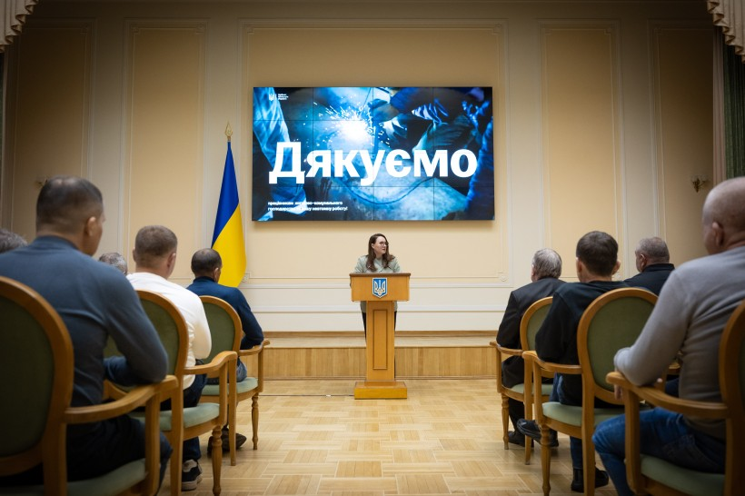 Жінка виступає за трибуною з гербом України в офіційній залі перед аудиторією. На великому екрані позаду неї напис «Дякуємо», а ліворуч стоїть державний прапор.