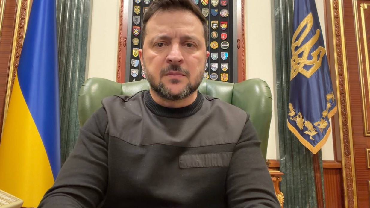 Чоловік у чорному светрі сидить у шкіряному кріслі та звертається на камеру. Позаду нього — Державний Прапор України та синій прапор із золотим тризубом, а на стіні розміщена колекція військових шевронів.