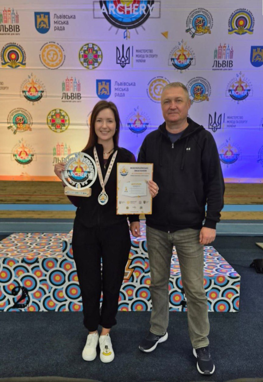 Нагородження спортсменки на змаганнях зі стрільби з лука у Львові. Дівчина з медаллю, грамотою та кубком стоїть поруч із чоловіком на фоні тематичного банера з гербами міста, міністерства та федерації.