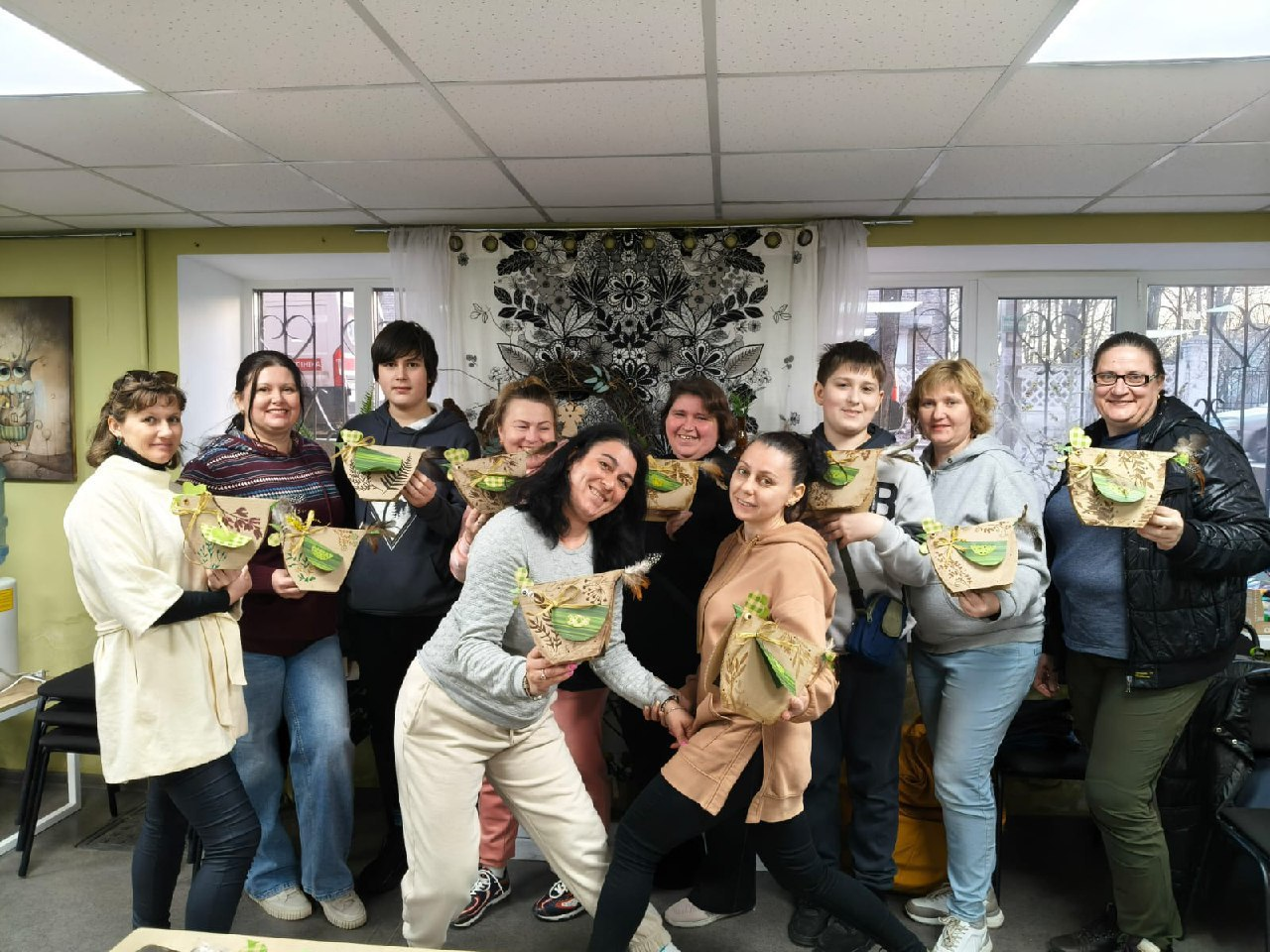 Група дорослих та підлітків позує у світлому приміщенні, тримаючи в руках власноруч виготовлені декоративні вироби у формі пташок. Учасники заходу посміхаються на фоні вікна та декоративного панно з рослинним орнаментом.