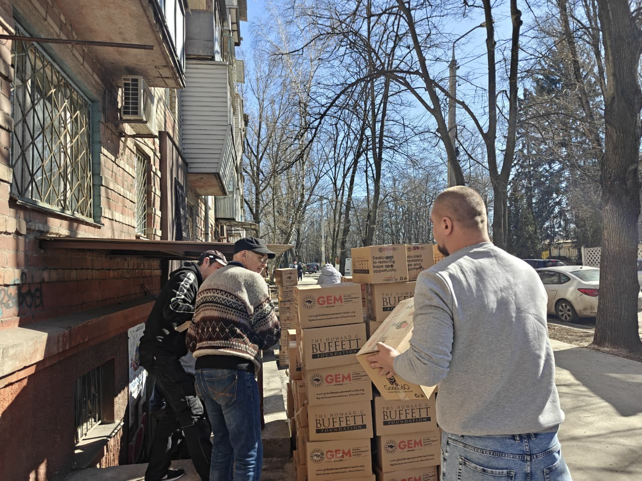 Чоловіки розвантажують коробки з гуманітарною допомогою від фондів GEM та Howard G. Buffett біля житлового будинку. Процес передачі допомоги відбувається на вулиці у сонячний день.