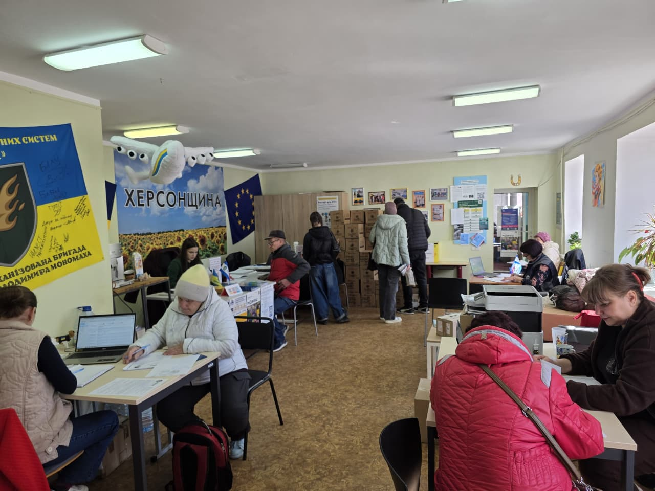 Робота гуманітарного офісу «Херсонщина»: відвідувачі отримують консультації за столами, а на задньому плані триває видача коробок із допомогою. Приміщення прикрашене прапорами та патріотичними плакатами.