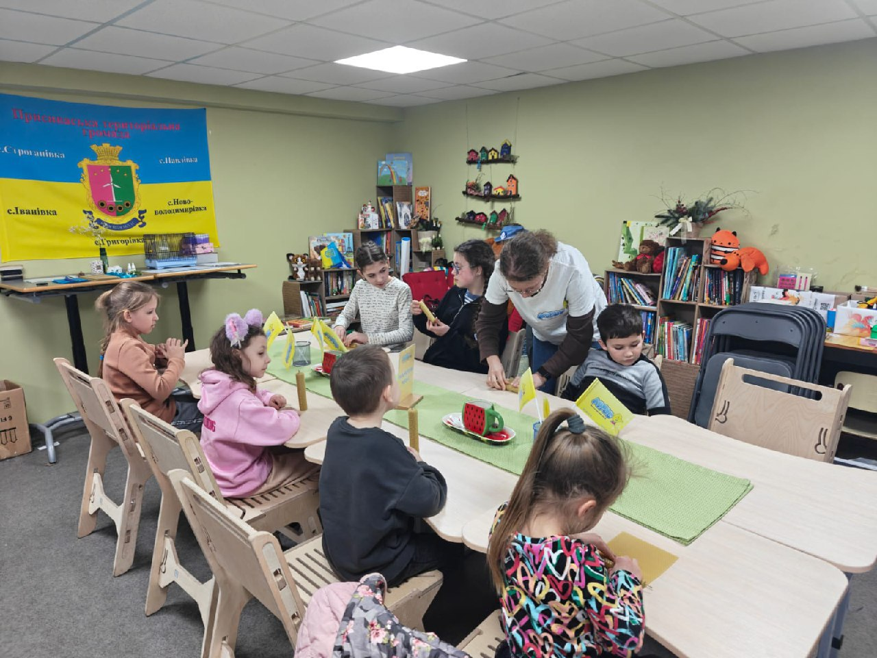 Група дітей та доросла жінка займаються творчістю за великим столом у дитячому куточку гуманітарного хабу. На задньому плані — прапор громади, полиці з іграшками та книжками.