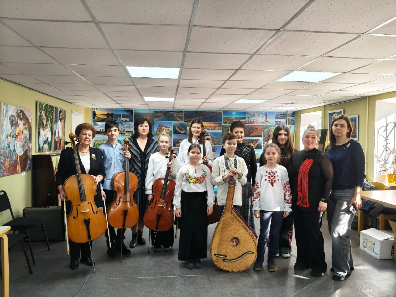 Група дорослих та дітей-музикантів позує в мистецькому залі з віолончелями та бандурою. На стінах приміщення розміщені картини та фотографії, що створюють творчу атмосферу під час культурного заходу.