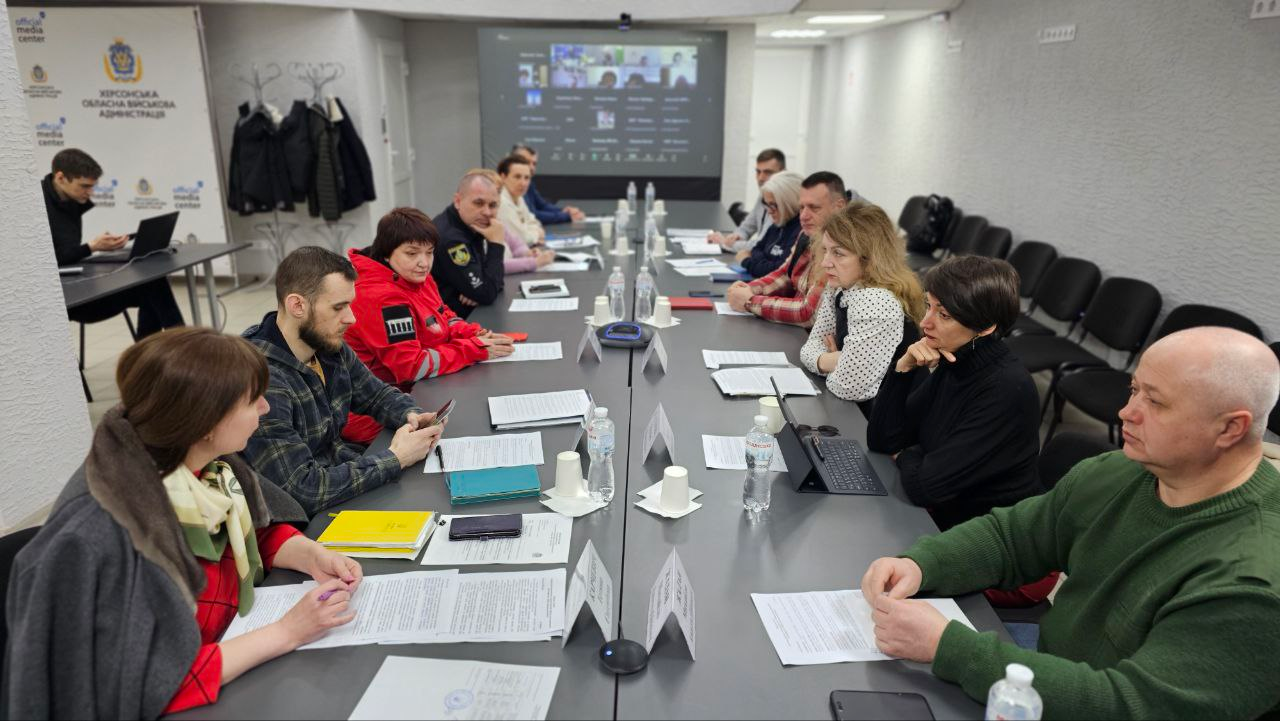 Учасники наради сидять за довгим столом у конференц-залі з документами та гаджетами. На фоні видно екран із відеозв'язком та банер Херсонської обласної державної адміністрації.