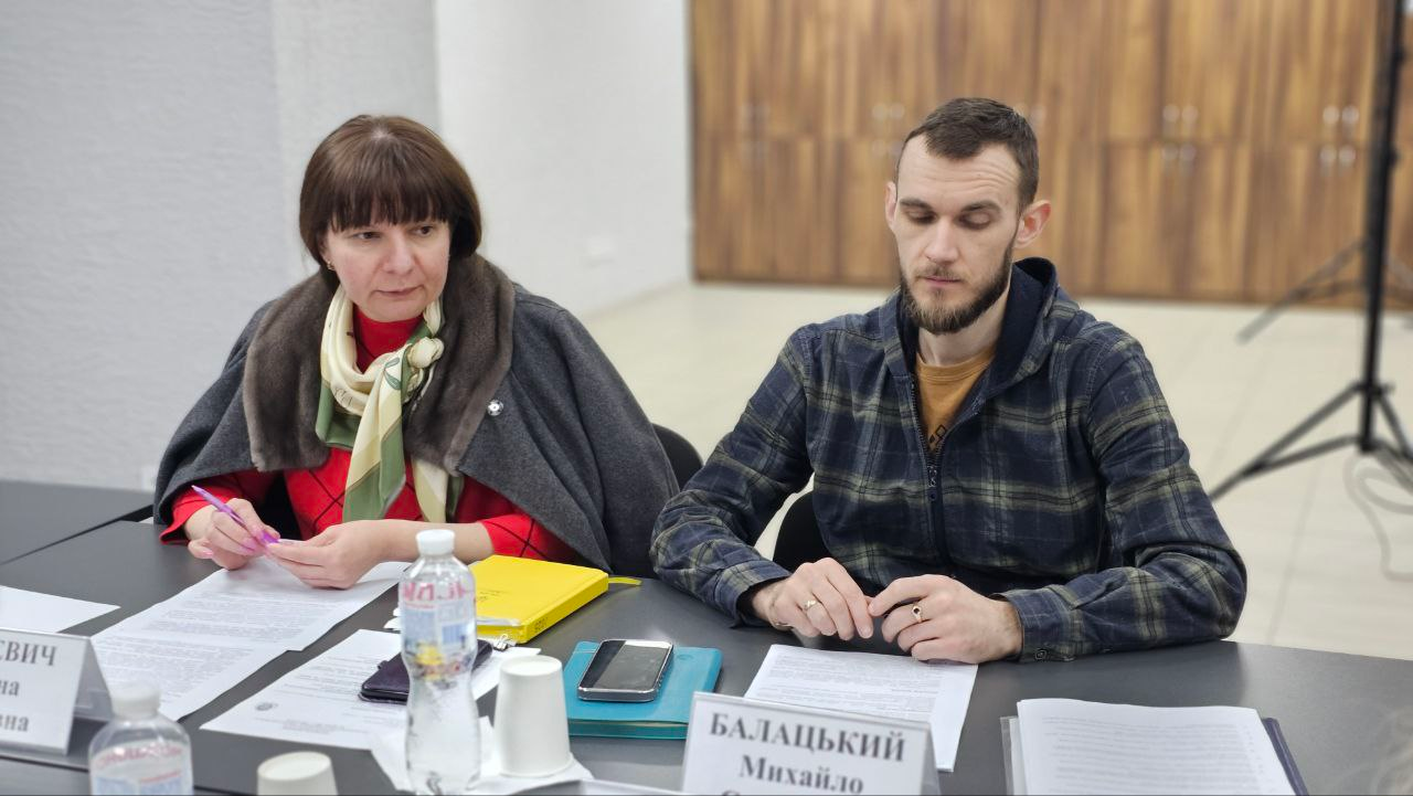 Жінка та чоловік сидять за робочим столом під час офіційного заходу або наради. Перед ними розкладені документи, блокноти, мобільні телефони та встановлені іменні таблички.