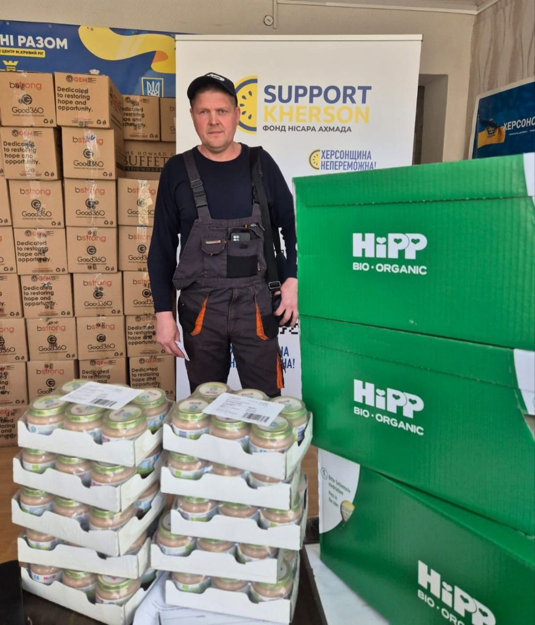Чоловік у робочому одязі стоїть поруч із великою кількістю коробок та упаковок дитячого харчування HiPP. На фоні видно банери «Support Kherson» та логотипи гуманітарних організацій, що свідчить про процес отримання або розподілу допомоги.