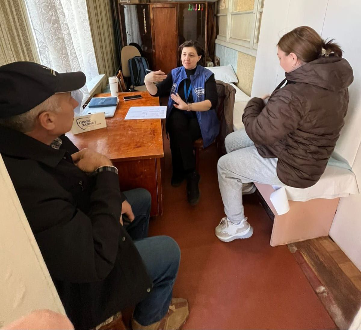 Процес надання безоплатної правової допомоги. Юристка у синьому жилеті з логотипом благодійного фонду консультує двох відвідувачів, сидячи за робочим столом із документами та табличкою «Право на захист».