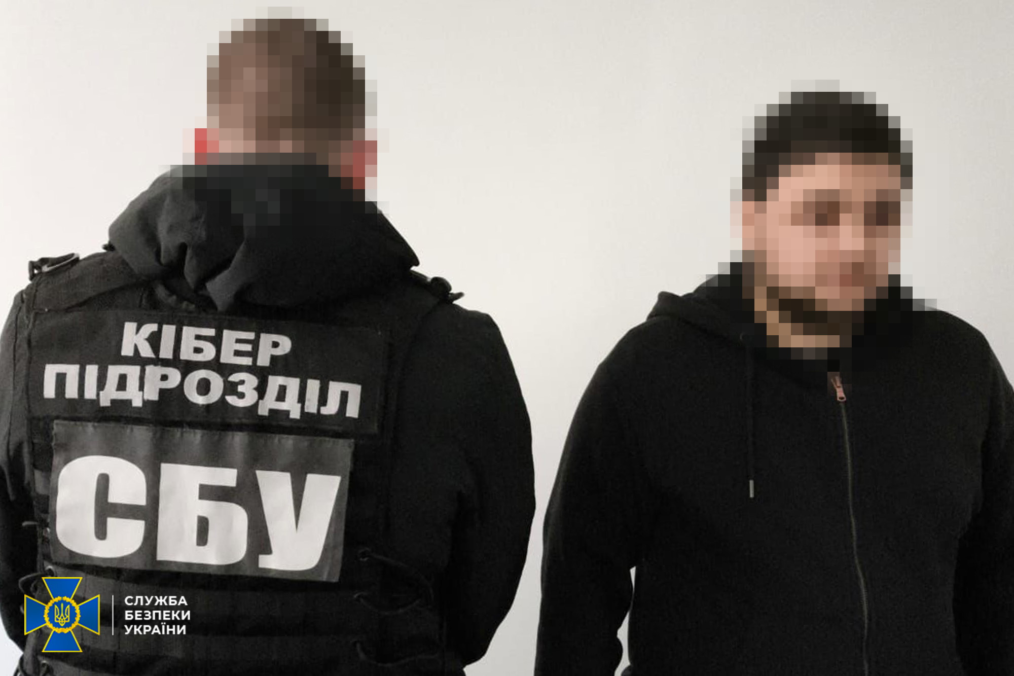 Представник кіберпідрозділу СБУ у тактичному спорядженні стоїть поруч із затриманим чоловіком на світлому фоні. Обличчя обох осіб заретільовані, а на спині офіцера чітко видно написи «КІБЕР ПІДРОЗДІЛ» та «СБУ».