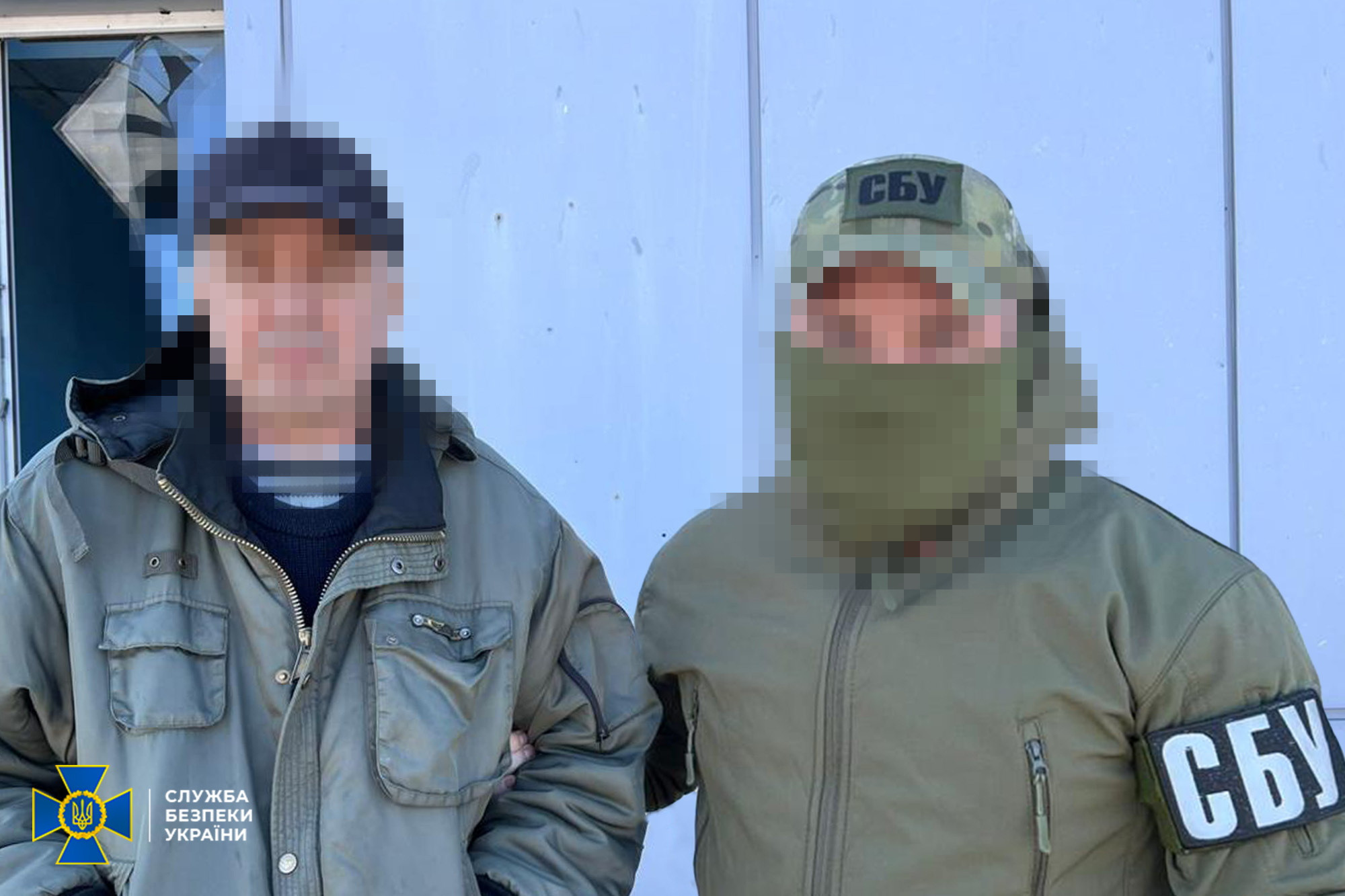 Співробітник СБУ у військовій формі та балаклаві тримає під руку затриманого чоловіка у цивільному одязі. Обличчя обох осіб заретільовані, а на рукаві та кепці офіцера чітко видно шеврони «СБУ».
