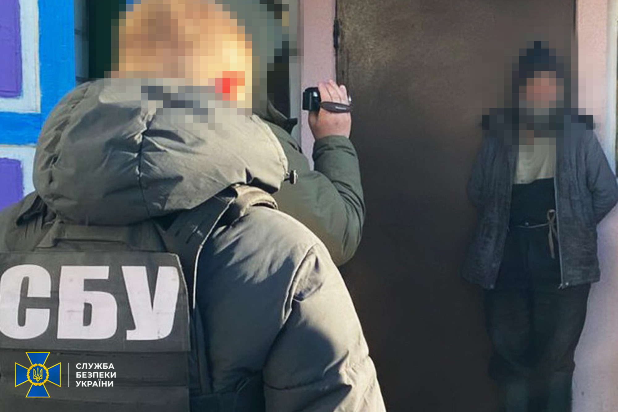 Співробітник СБУ у зимовій куртці з великим написом на спині веде відеофільмування чоловіка у цивільному одязі, який стоїть біля входу до приміщення. Обличчя обох осіб заретільовані, у лівому нижньому куті розміщено офіційний логотип Служби безпеки України.