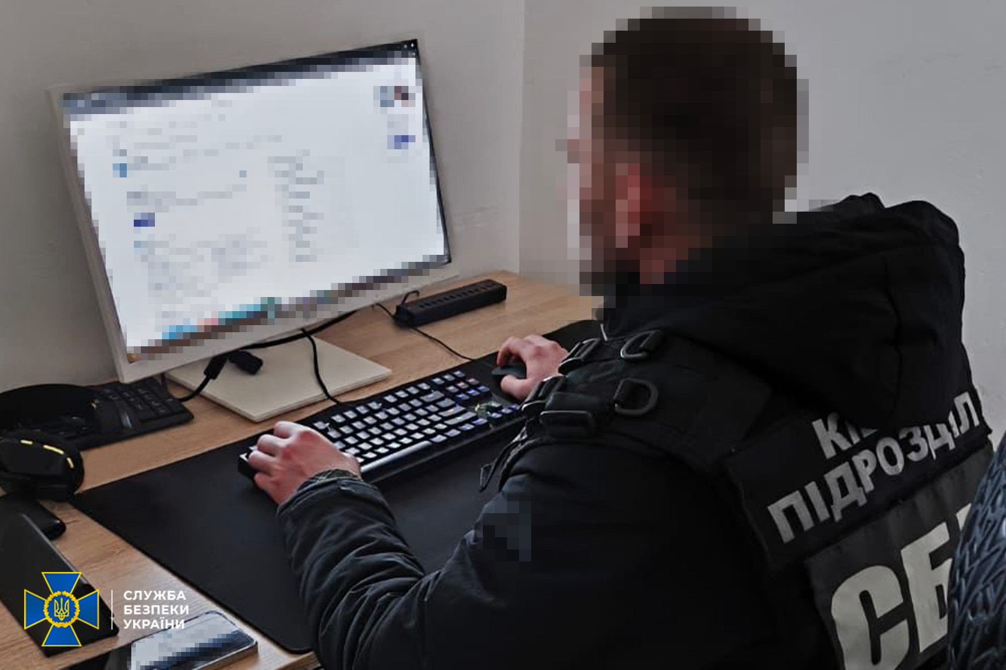 Співробітник кіберпідрозділу СБУ у тактичному спорядженні працює за комп'ютером. Обличчя особи та зображення на моніторі заретільовані, на спині офіцера видно напис «КІБЕР ПІДРОЗДІЛ СБУ», у кутку — офіційний логотип служби.