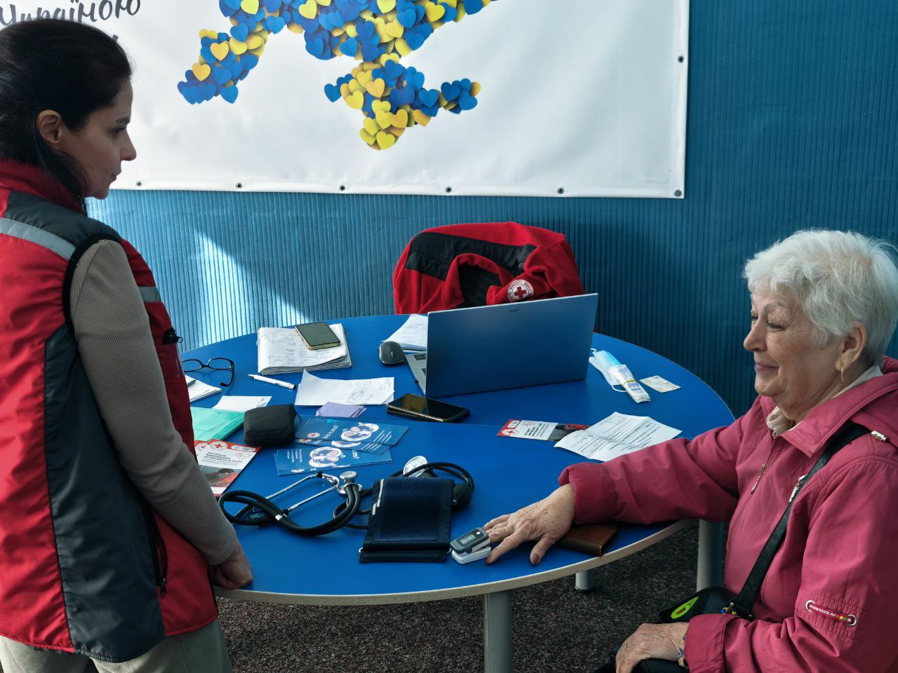 Процес надання медичної допомоги літній жінці волонтеркою Червоного Хреста. На синьому столі розкладено тонометр, стетоскоп, пульсоксиметр та ноутбук; на задньому плані — патріотичне панно з мапою України з синьо-жовтих сердець.