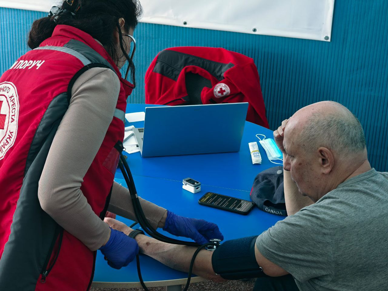 Волонтерка Товариства Червоного Хреста України вимірює артеріальний тиск літньому чоловіку за допомогою тонометра. На столі поруч із пацієнтом лежать ноутбук, пульсоксиметр та медична маска.