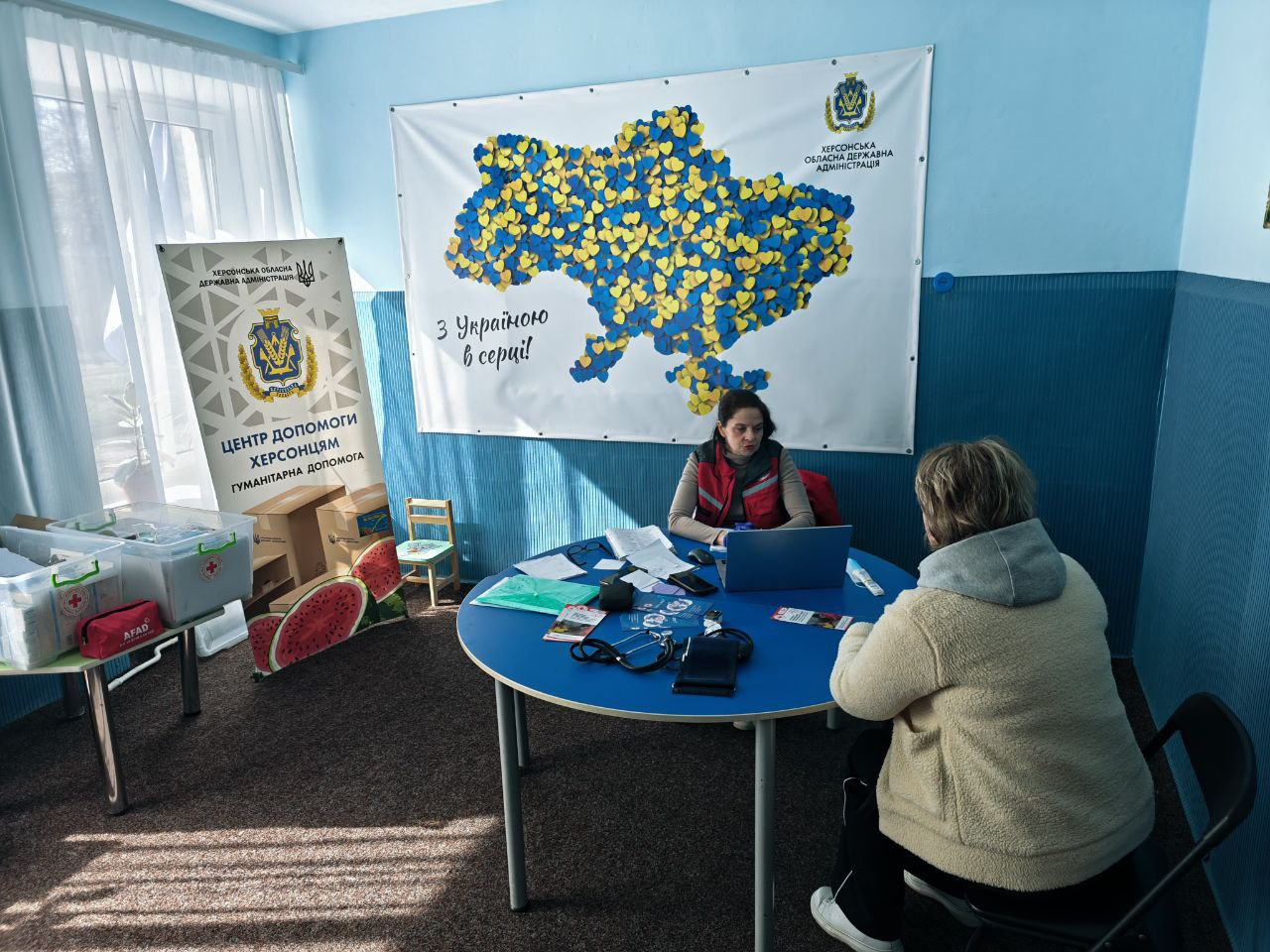 Надання медичної допомоги представницею Червоного Хреста жінці у приміщенні гуманітарного хабу. На задньому плані — велике панно з мапою України із синьо-жовтих сердець, інформаційний банер про гуманітарну допомогу та бокси з медичним приладдям.