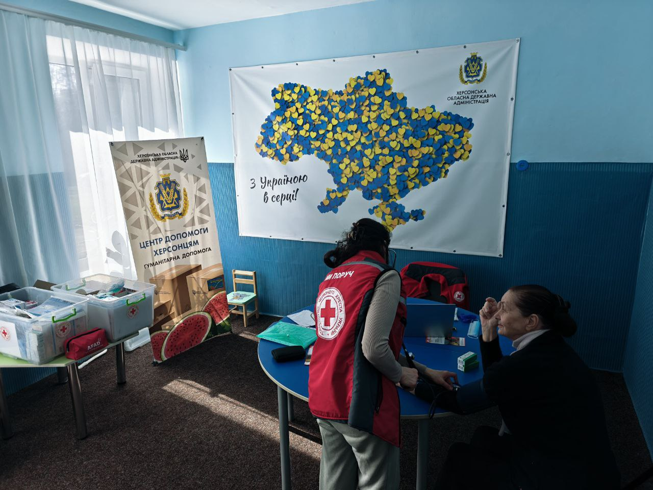 Надання медичної допомоги представницею Червоного Хреста жінці у приміщенні гуманітарного хабу. На задньому плані — велике панно з мапою України із синьо-жовтих сердець, інформаційний банер про гуманітарну допомогу та бокси з медичними приладдями.