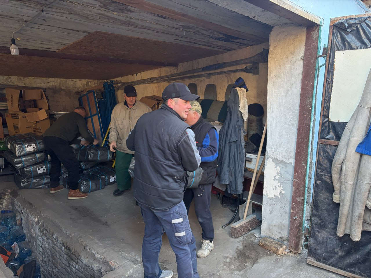 Чоловіки розвантажують та складають пакунки з гуманітарною або технічною допомогою у складському приміщенні. На задньому плані видно картонні коробки та інше обладнання.