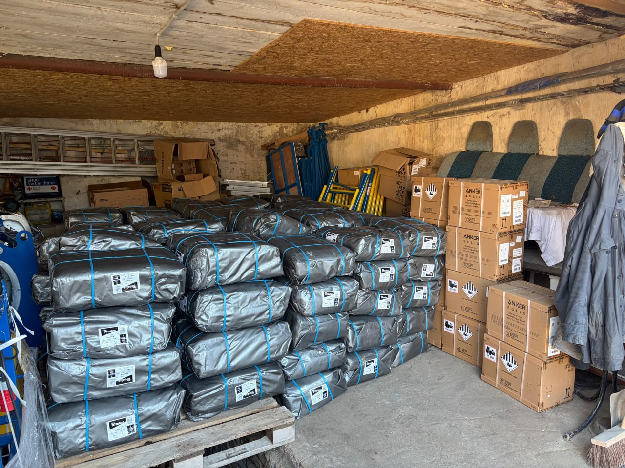 Складське приміщення, заповнене гуманітарною або технічною допомогою. У центрі складені штабелями пакунки у сірій захисній плівці, перев'язані синіми стрічками, а праворуч — картонні коробки з маркуванням виробника Anker SOLIX.