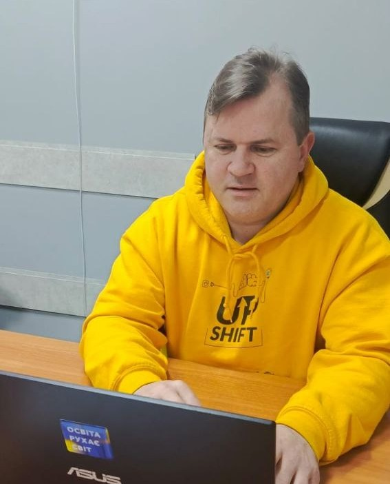 Чоловік у яскраво-жовтому худі з логотипом молодіжної програми «UPSHIFT» працює за ноутбуком ASUS. На ноутбуці наклеєно стікер із патріотичним гаслом «Освіта рухає світ».