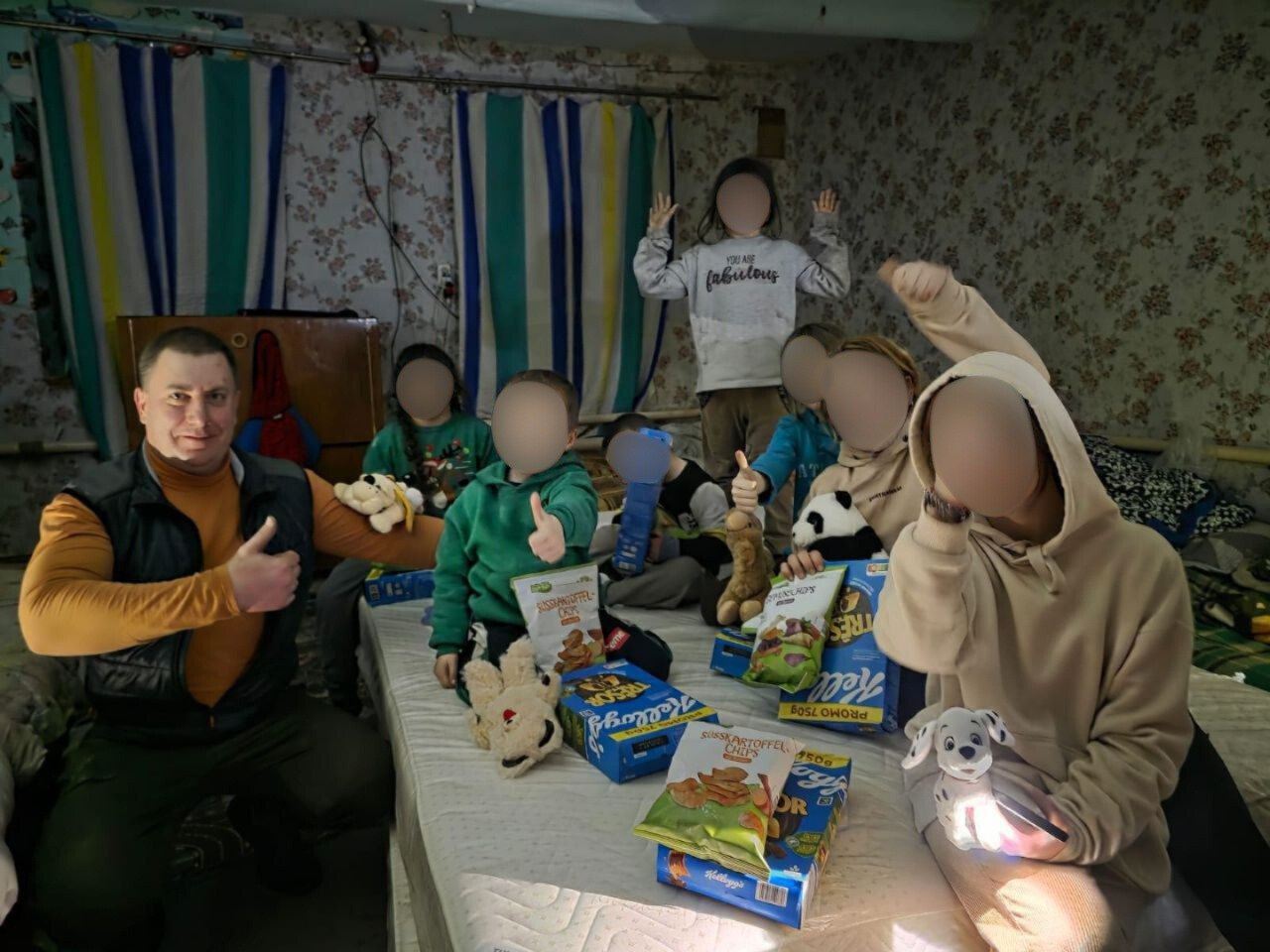 Група дітей та дорослий чоловік радісно позують у кімнаті, оточені пакунками з гуманітарною допомогою. Діти тримають у руках м'які іграшки та пачки зі снеками, демонструючи жест «клас».