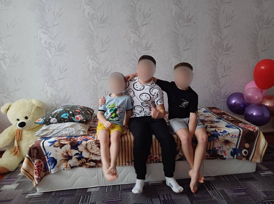 Троє хлопчиків сидять на ліжку в домашній кімнаті, обійнявши один одного. Поруч із ліжком стоїть великий іграшковий ведмідь, а з іншого боку — різнокольорові повітряні кульки.