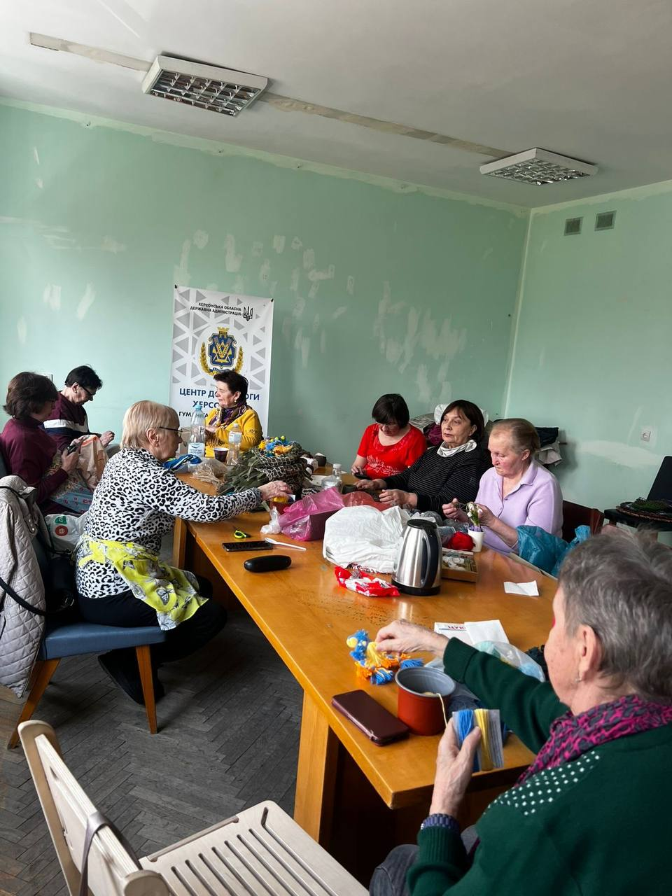 Група жінок займається рукоділлям за великим столом у гуманітарному хабі. На фоні видно інформаційний банер «Центр допомоги херсонцям» з офіційною символікою.