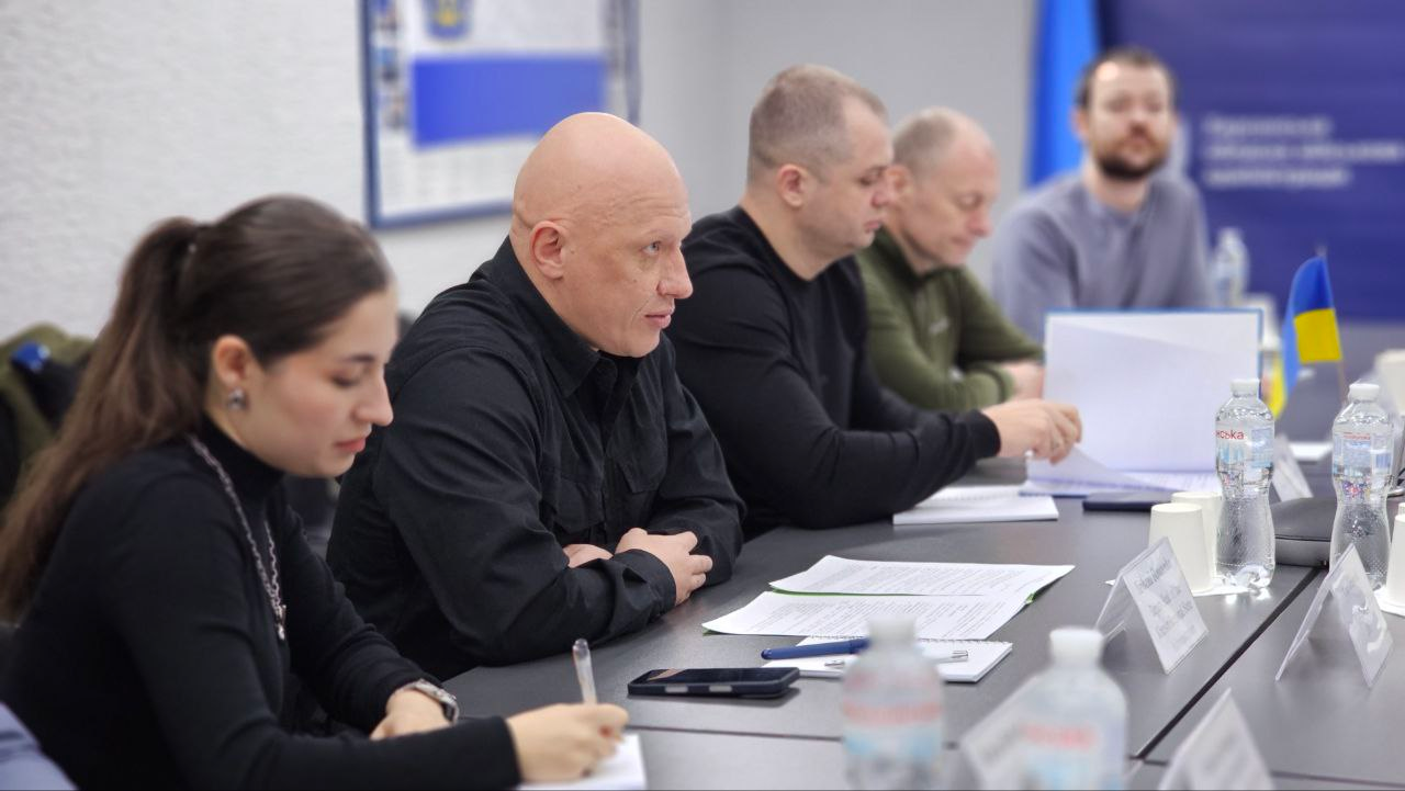 Учасники офіційної наради або засідання за довгим робочим столом. На передньому плані жінка занотовує інформацію, поруч — троє чоловіків у темному одязі та військовій формі вивчають документи. На столі розміщені пляшки з водою, папери та іменні таблички, на задньому плані — державний прапор України та офіційний банер.