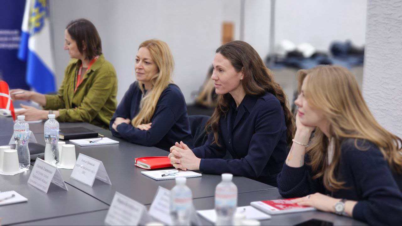 Чотири жінки за столом під час офіційної зустрічі з міжнародними партнерами. Учасниці уважно слухають виступи, на столі розкладено блокноти, воду та іменні таблички. Поруч стоїть прапорець Данії.