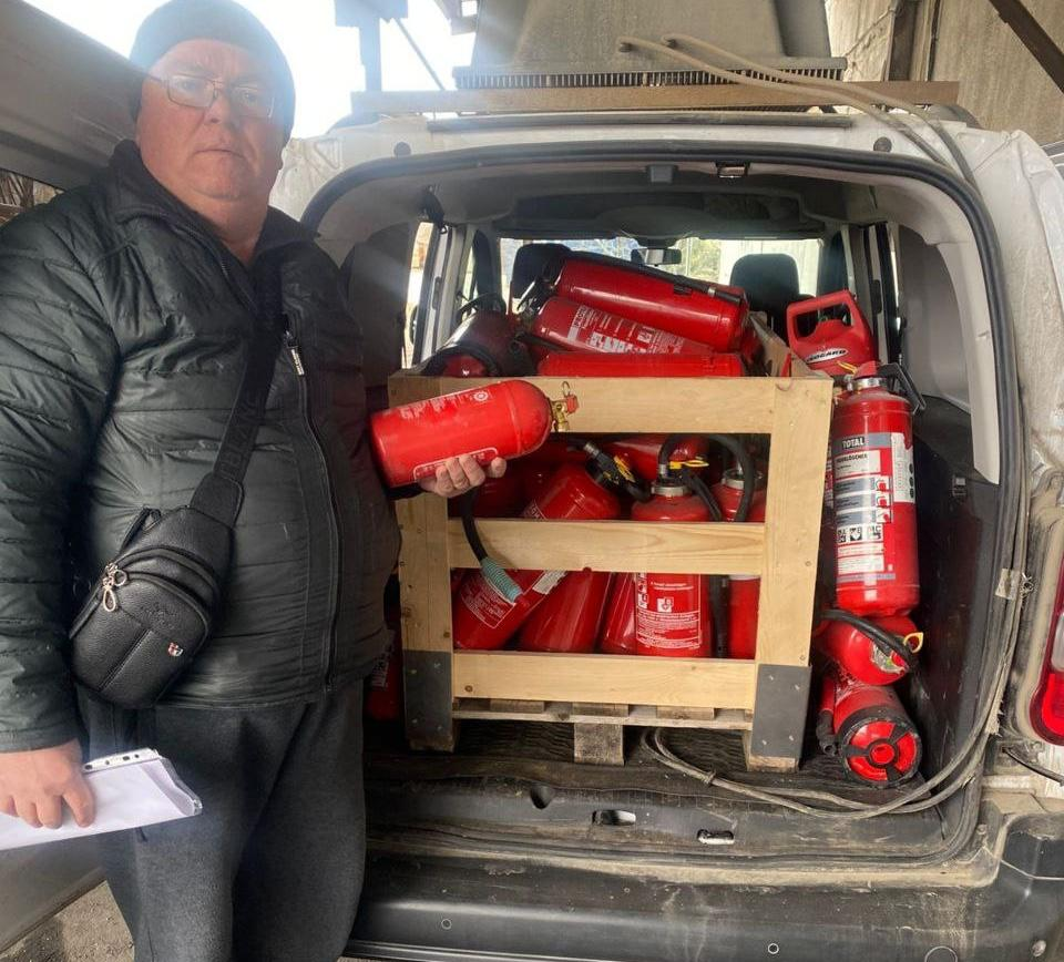 Чоловік тримає вогнегасник біля відкритого багажника автомобіля, вщент заповненого червоними вогнегасниками в дерев'яному каркасі.