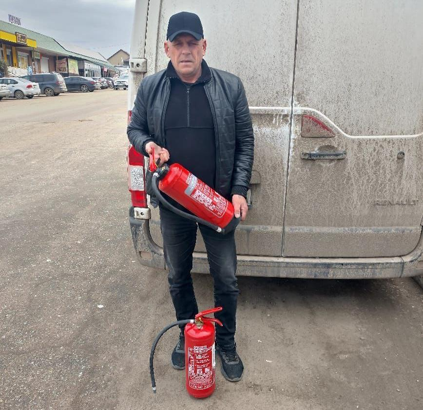 Чоловік у темному одязі стоїть біля задніх дверей мікроавтобуса, тримаючи в руках вогнегасник. Ще один вогнегасник стоїть на землі поруч із ним.
