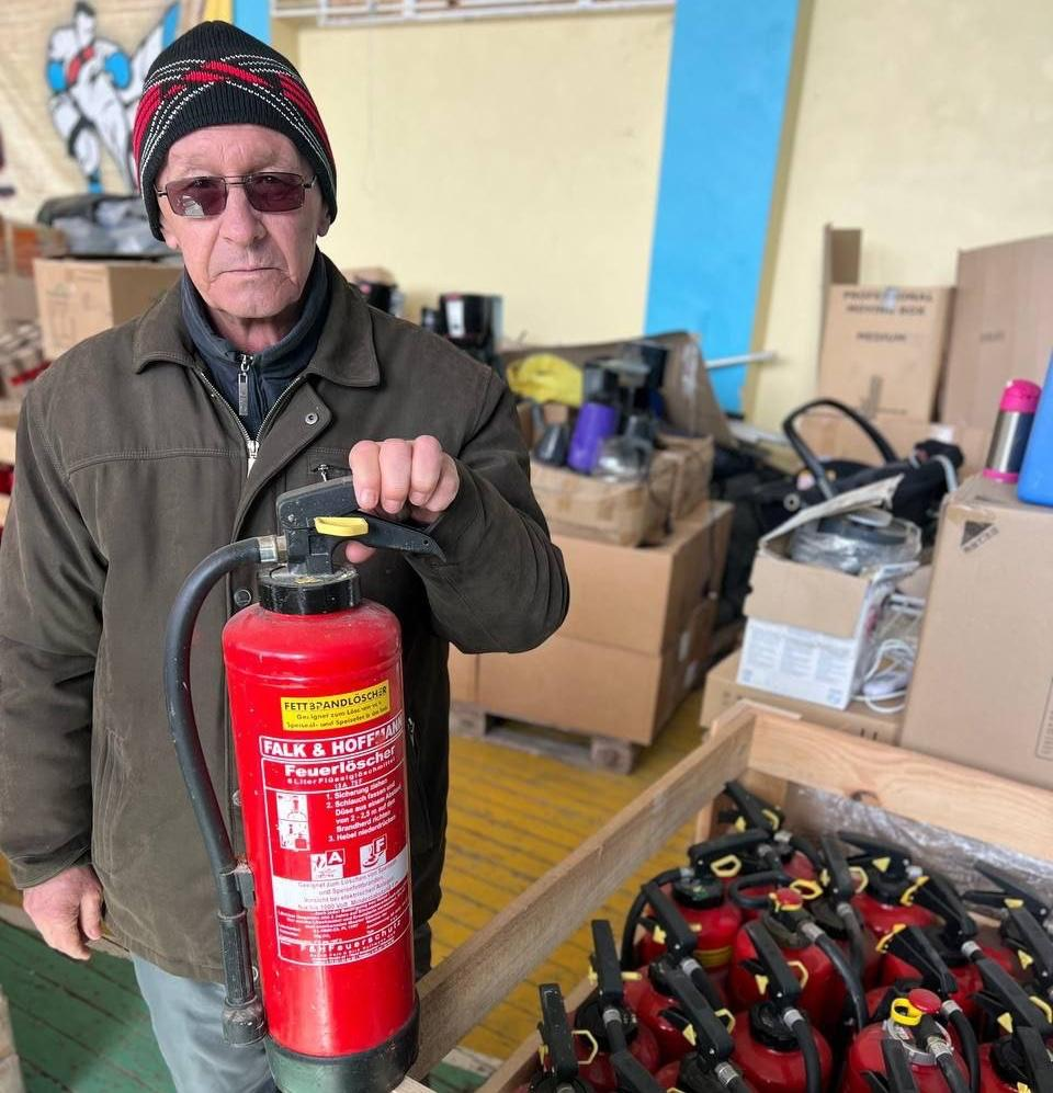 Чоловік у куртці та шапці тримає червоний вогнегасник на фоні складу з коробками та іншими вогнегасниками.