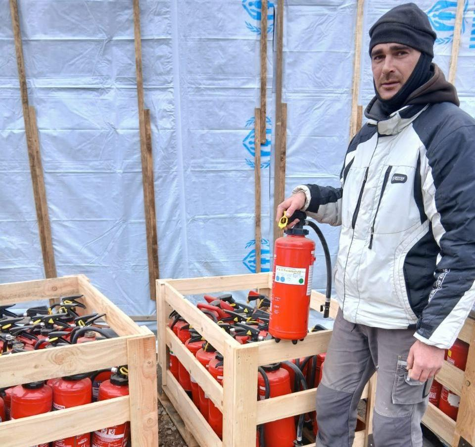 Чоловік у зимовому одязі тримає червоний вогнегасник поруч із дерев'яними ящиками, що вщент заповнені вогнегасниками.