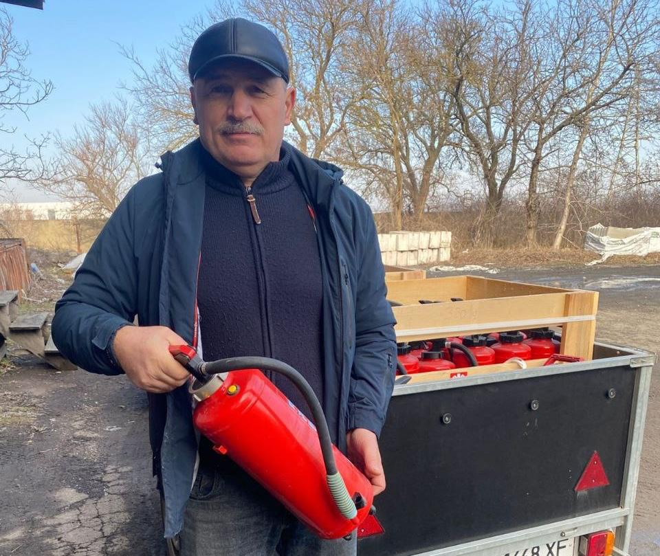 Чоловік тримає в руках вогнегасник на фоні відкритого автомобільного причепа, заповненого червоними вогнегасниками в дерев'яному ящику.