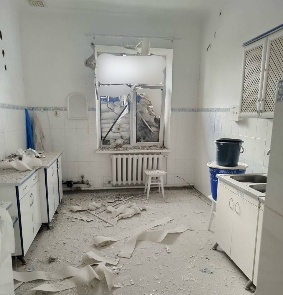 Пошкоджена кухня після обстрілу: розбите вікно закладене мішками з піском, на підлозі розкидане сміття, уламки та шматки шпалер, меблі зміщені.