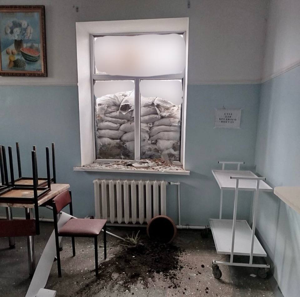 Пошкоджений обстрілом кабінет закладу охорони здоров'я. Вікно закладене мішками з піском, на підлозі розбитий горщик із землею, меблі та речі розкидані.