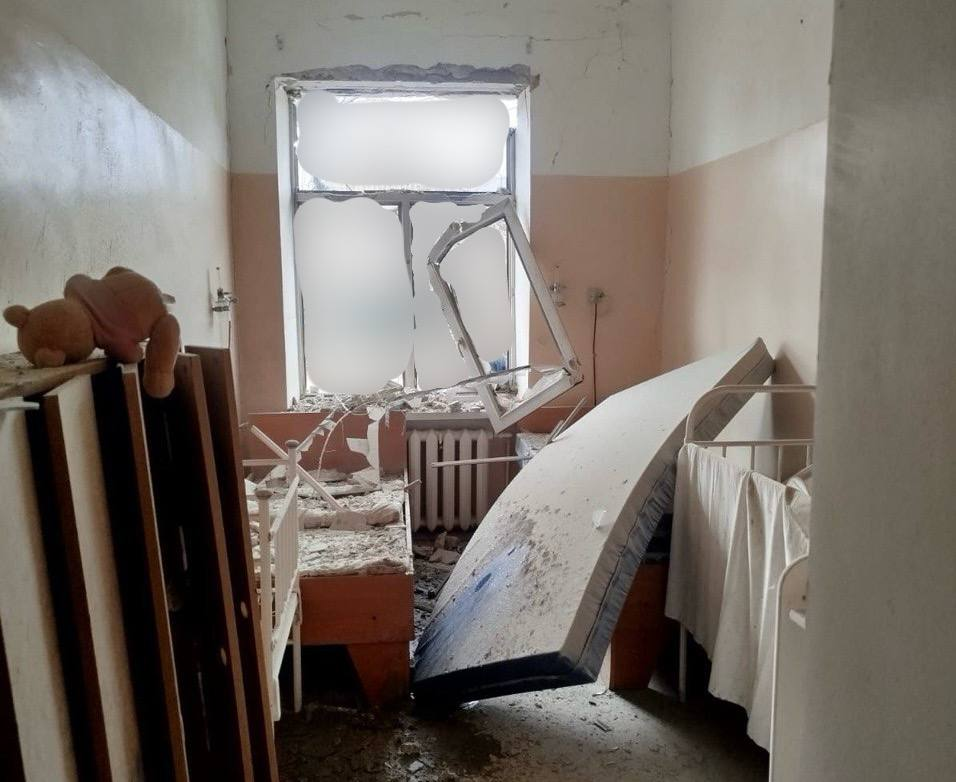 Зруйнована дитяча кімната або палата після обстрілу. Вибите вікно, перекинутий матрац, пошкоджені ліжка та м’яка іграшка ведмідь на уламках.