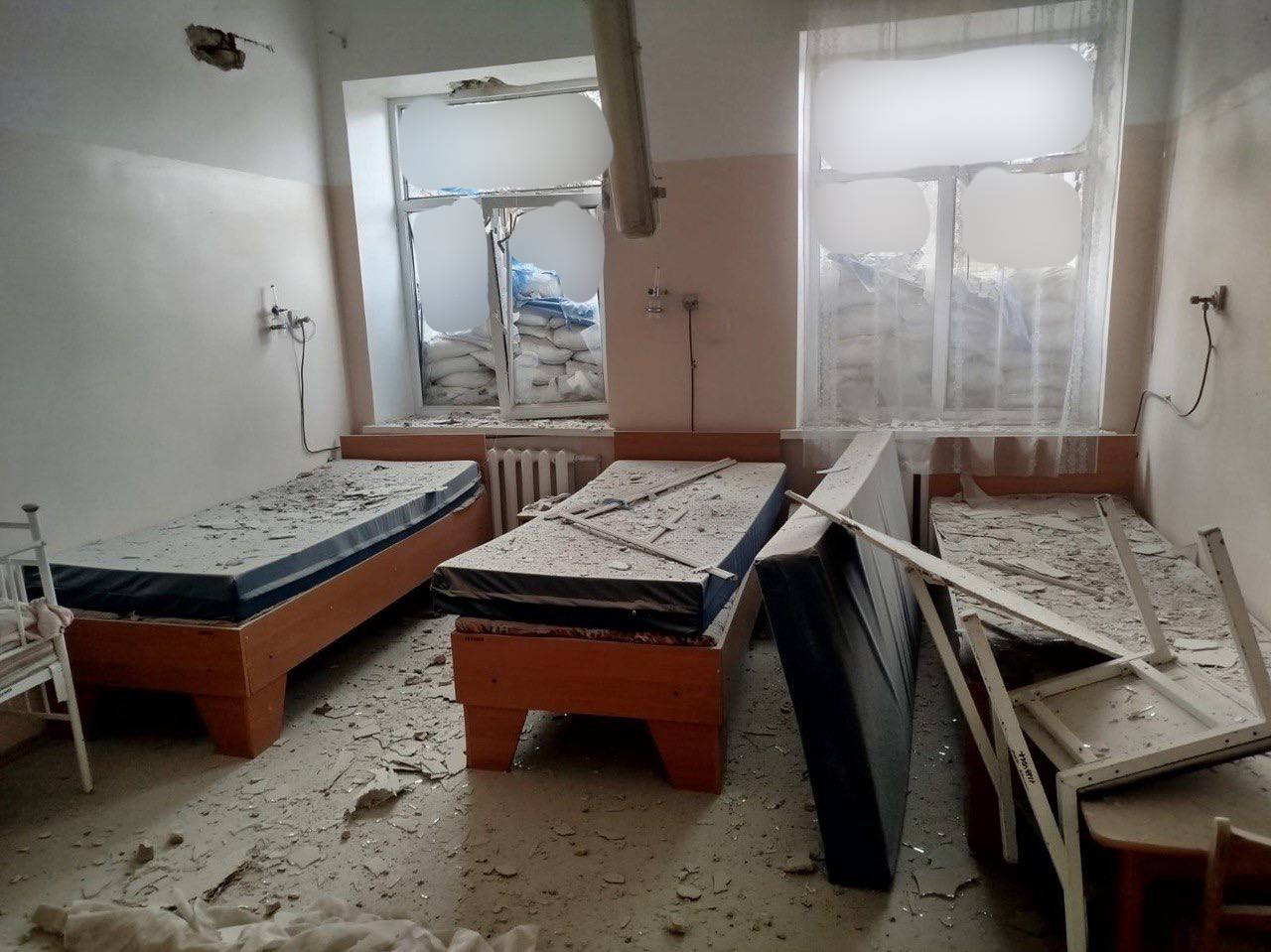 Зруйноване приміщення з кількома ліжками, які вкриті будівельним сміттям та уламками. Вікна пошкоджені й закладені мішками з піском, меблі перекинуті.
