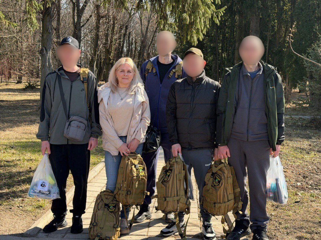 Група людей на прогулянці в парку сонячного дня. Чоловіки та жінка позують для фото, тримаючи в руках тактичні рюкзаки піщаного кольору та пакети з речами. Обличчя чоловіків приховані.