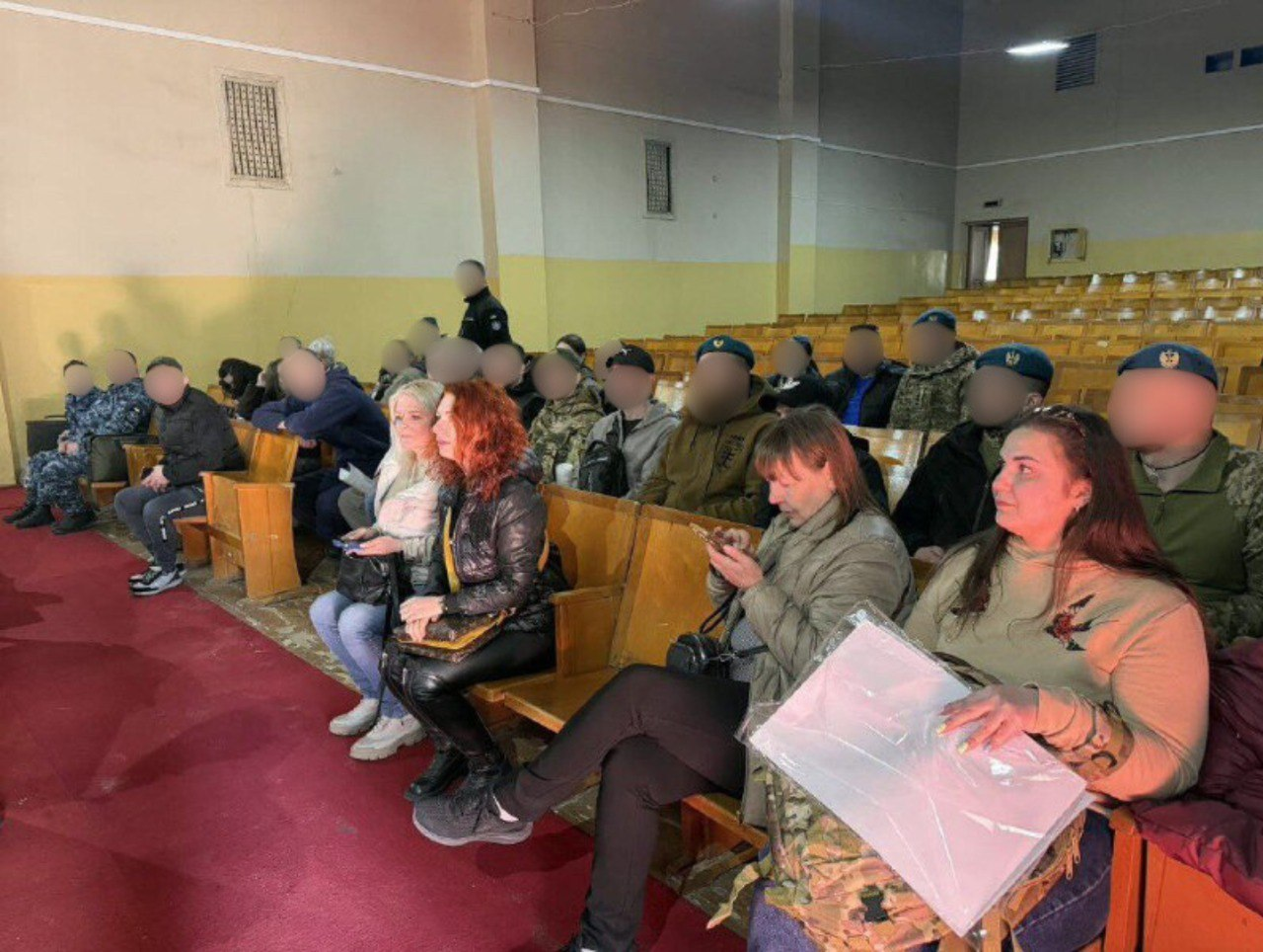 Група людей у залі для глядачів сидить на дерев'яних кріслах. У першому ряду жінки тримають у руках особисті речі та документи, деякі з них дивляться в мобільні телефони. На задніх рядах присутні люди у військовій та цивільній формі; обличчя всіх осіб на фото, крім жінок на першому ряду, приховані.