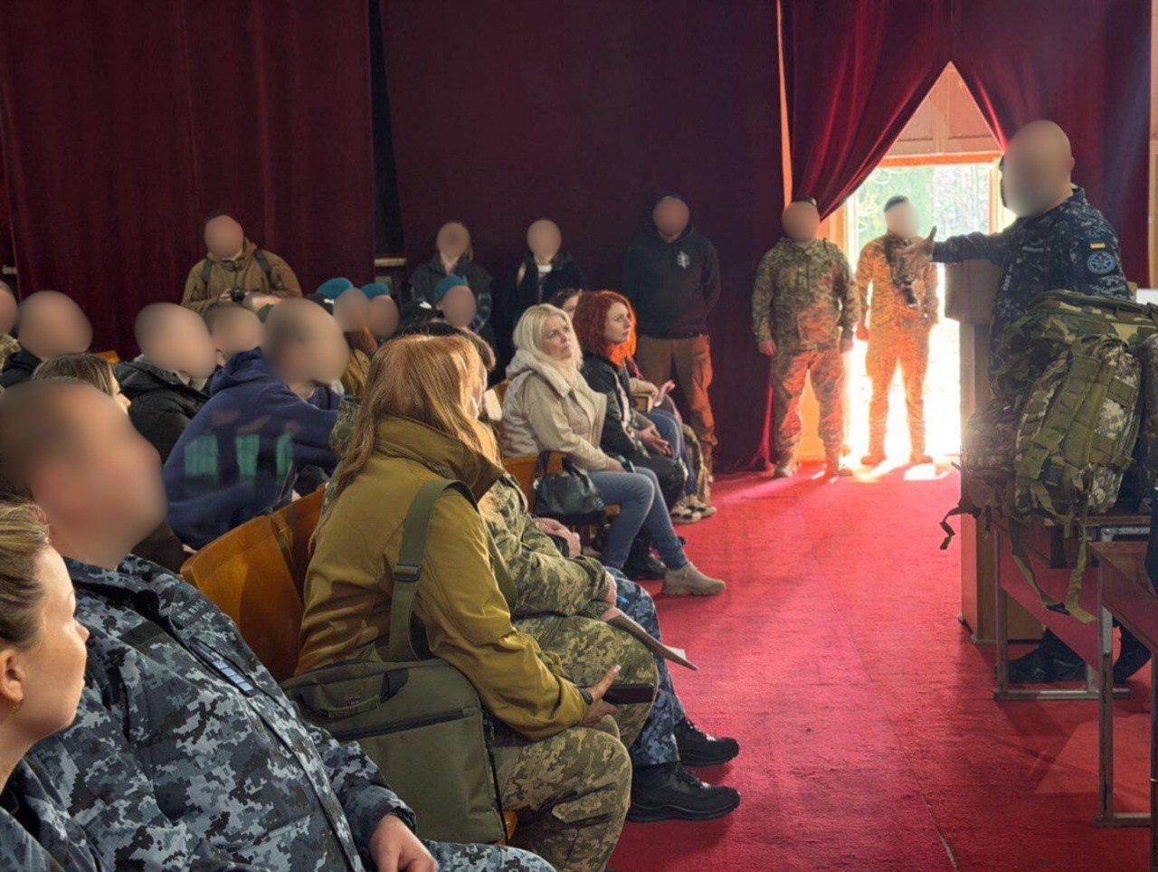 Зустріч у приміщенні з червоним килимовим покриттям та завісами. Чоловік у формі звертається до аудиторії, що складається з людей у військовій та цивільній формі, які сидять на дерев'яних кріслах. Обличчя присутніх, за виключенням декількох жінок на першому ряду, приховані.